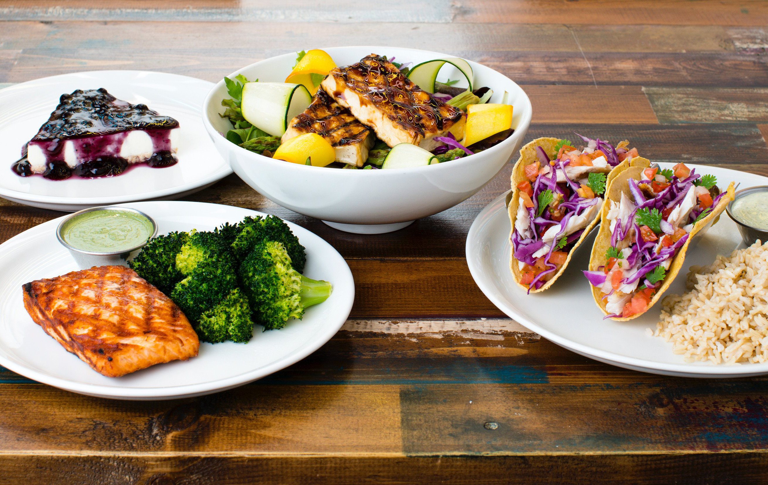 A range from the menu including their tacos, grilled salmon, tofu salad and blueberry cheesecake laid out on a wooden table