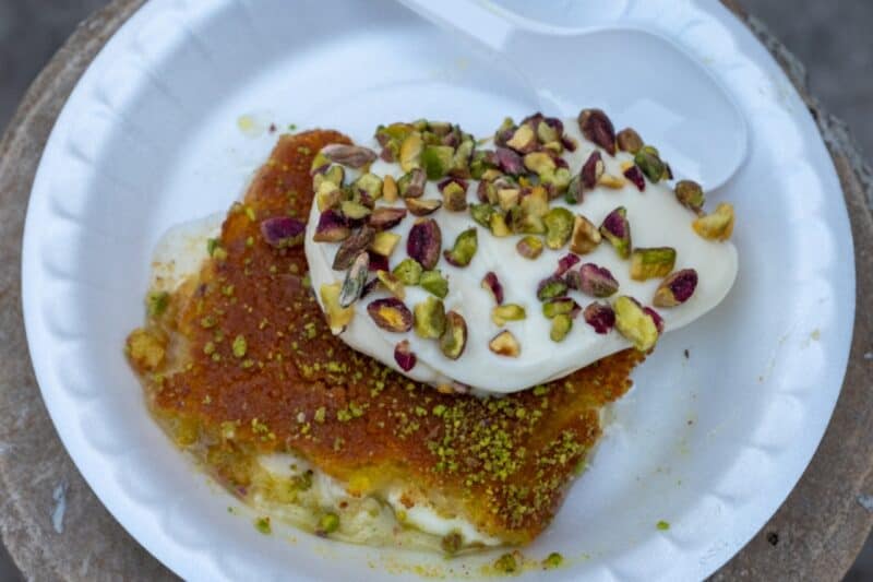Piece of kunafa served with cream on top and pistachios in Dubai on a disposable plate with a plastic spoon