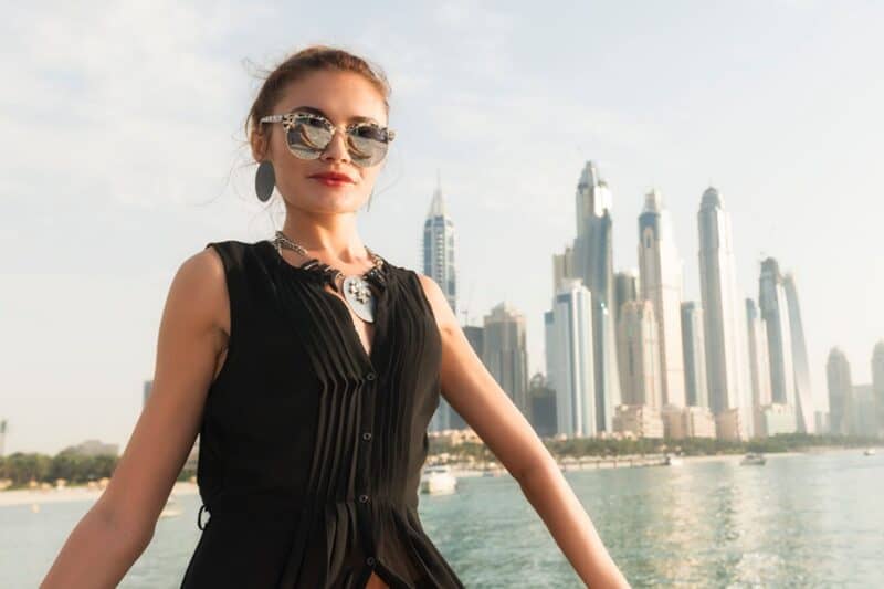 A woman sat on a yacht with Dubai Marina in the background, wearing a black top with her arms showing and sunglasses