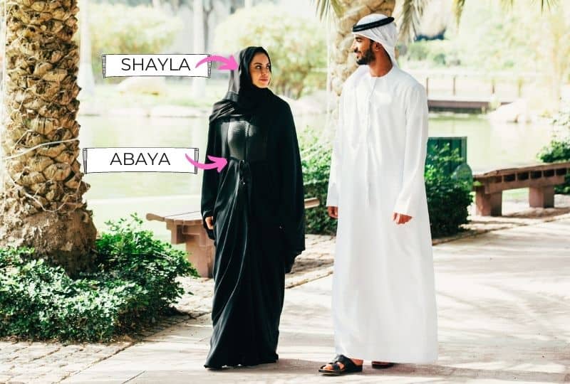Emirati man and emirati woman wearing tradtional Emirati outfits, the woman is labelled with the Emirati arabic names of the clothes. Shayla and Abaya. The shayla goes on the head of the woman to create hijab and the abaya is a clock over the body.