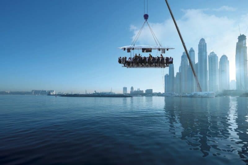 A dining table being hoisted by a crane over the sea with a view of the Dubai Marina skyline in the background. A unique bucketlist item in Dubai called Dinner in the Sky, literally!