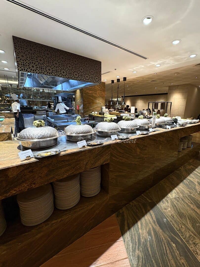 Various dishes inside silver pots with large decorative silver lids on a brown marble buffet counter with chefs preparing dishes in the background and plates ready for customers to pick up at JW Marriott Marquis, restaurant Kitchen 6