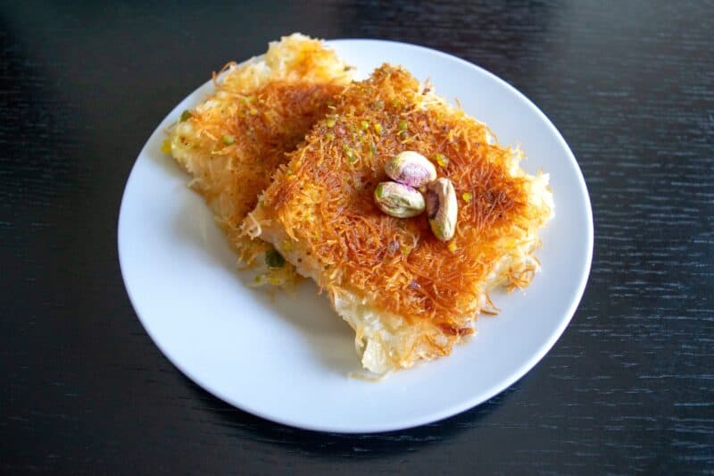 Two pieces of crunchy style kunafa on a ceramic plate with pistachio on top on a black table
