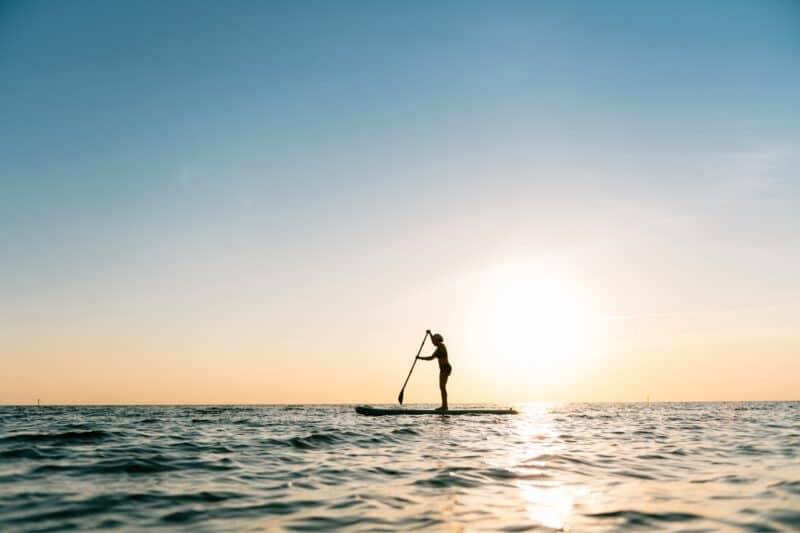 A woman standing on a paddle board reaching out with her paddle with the sun setting in the background on the sea