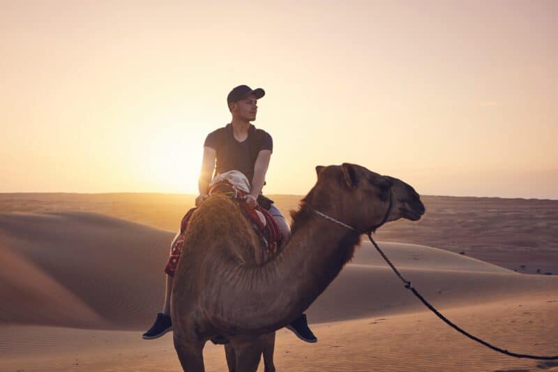 A man with a cap on riding a camel in the desert