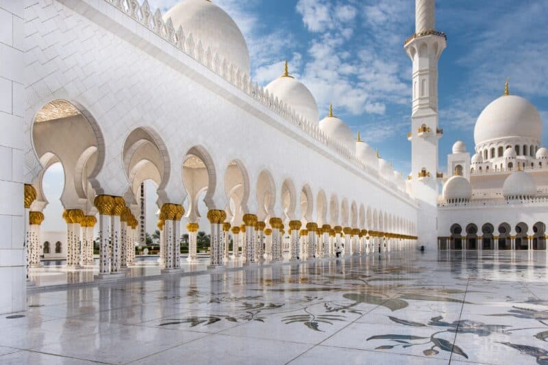 The courtyard area of Sheikh Zayed Grand Mosque in Abu Dhabi with a blue sky and clouds and the white marble domes.