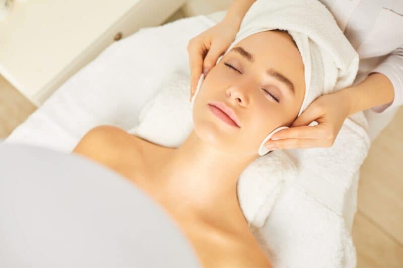 Woman lying down with a towel wrapped around her head as a lady gives her a facial with two cotton pads