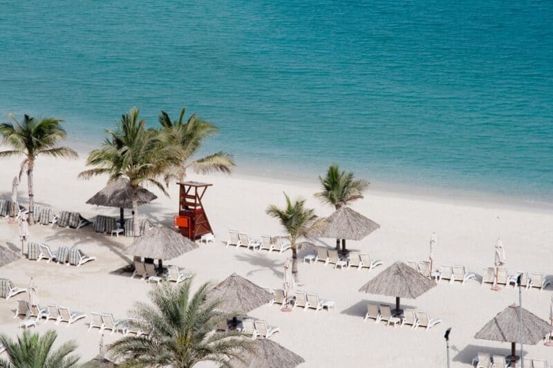 A beach resort in the UAE with pristine white beaches and lovely blue sea. White sun-loungers with tropical looking straw umbrelleas and palm trees
