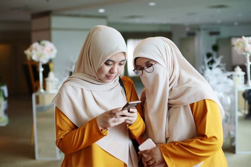 Two Muslim women looking at their phone and replying to messages from other sisters. Communication online can help with loneliness in Islam.