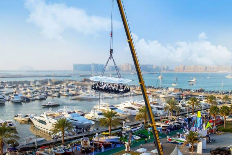 A dining table with people sat eating being hoisted by a crane over Dubai Marina with a view of Palm Jumeirah and Dubai Marina dock in the background called Dinner in the Sky.