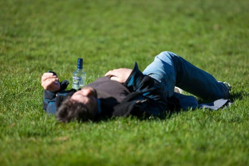A man passed out on the grass with a bottle of vodka next to him, clearly drunk in public