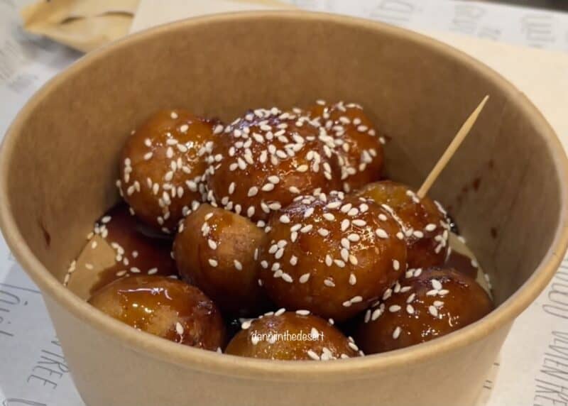 Gluten free Emirati Lugaimat served in a paper bowl with a toothpick, covered in date syrup and sesame seeds