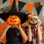 Two children dressed up for halloween with halloween decorations in the background and holding up pumpkins over their faces