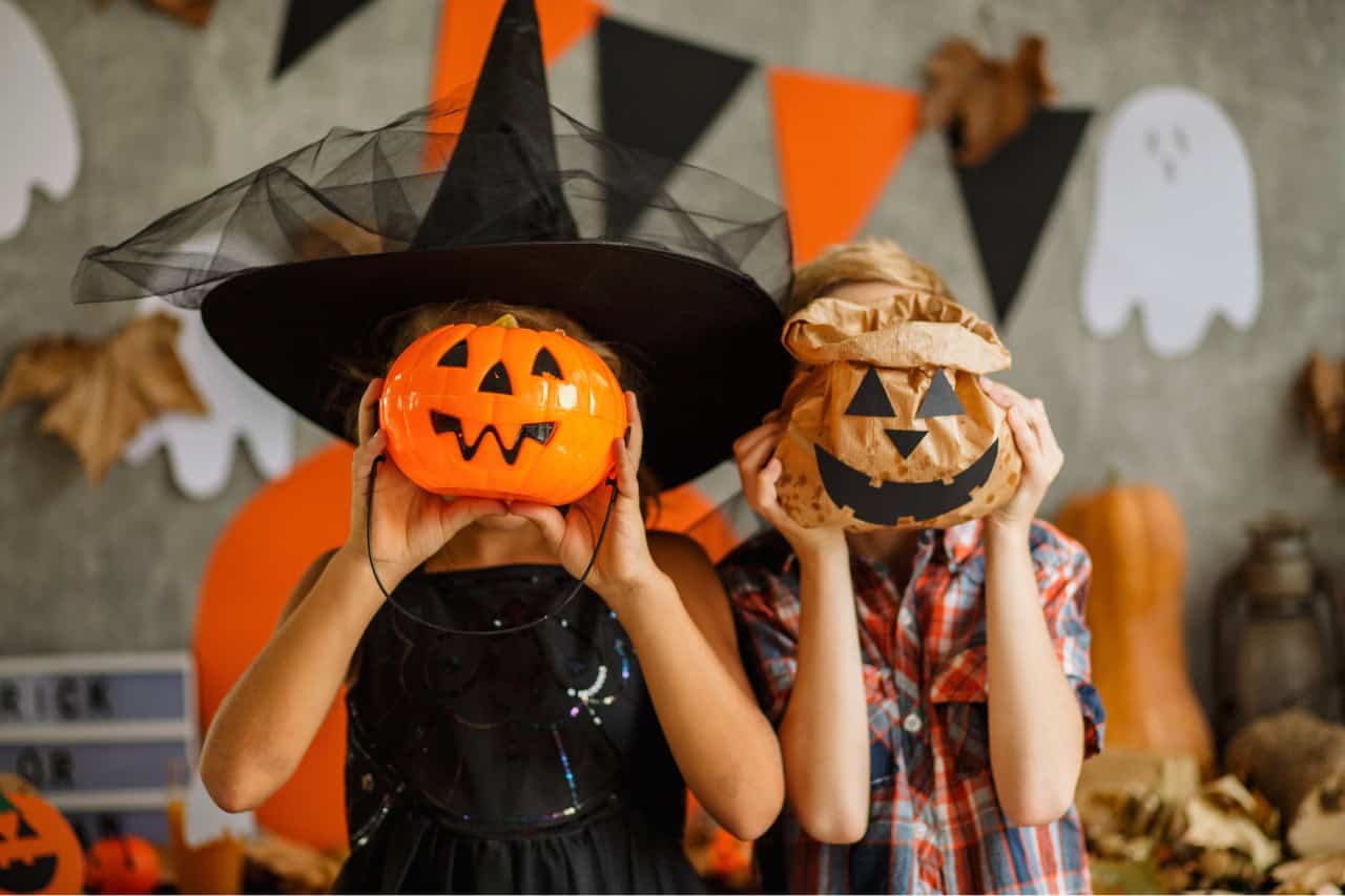 Two children dressed up for halloween with halloween decorations in the background and holding up pumpkins over their faces