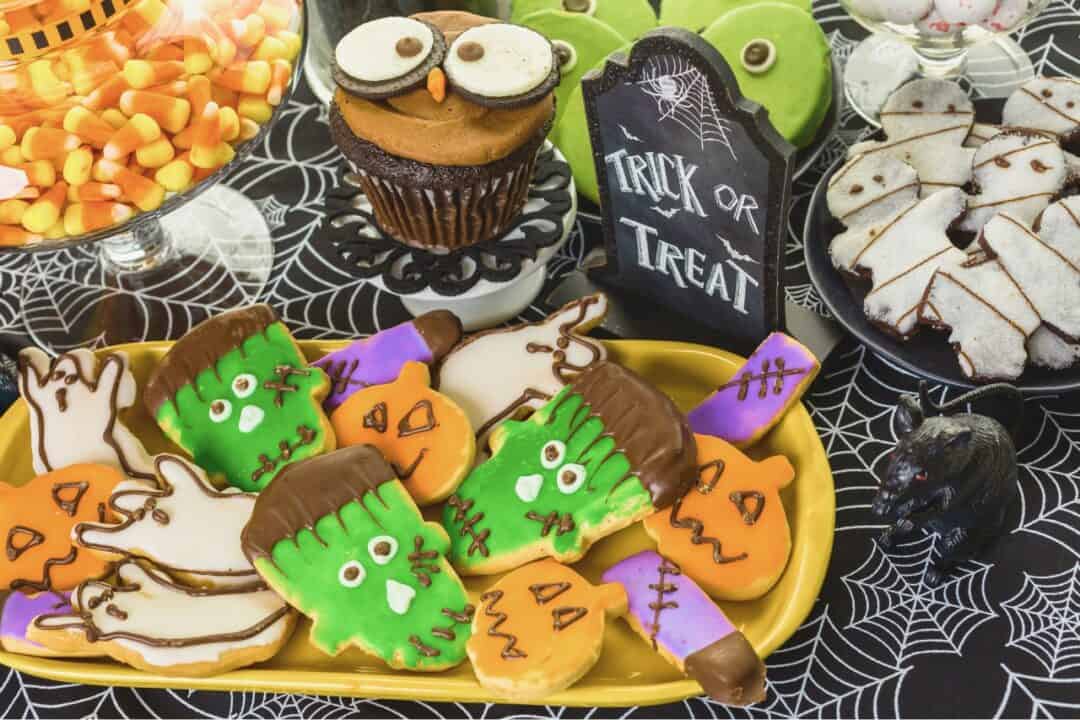 Different halloween treats for kids laid out on a spider wed table cloth with a sign that says trick or treat, including cookies, cup cakes and candy corn