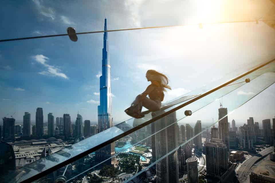 A woman sliding down the glass slide with a view of the Burj Khalifa in the background and the sun shining