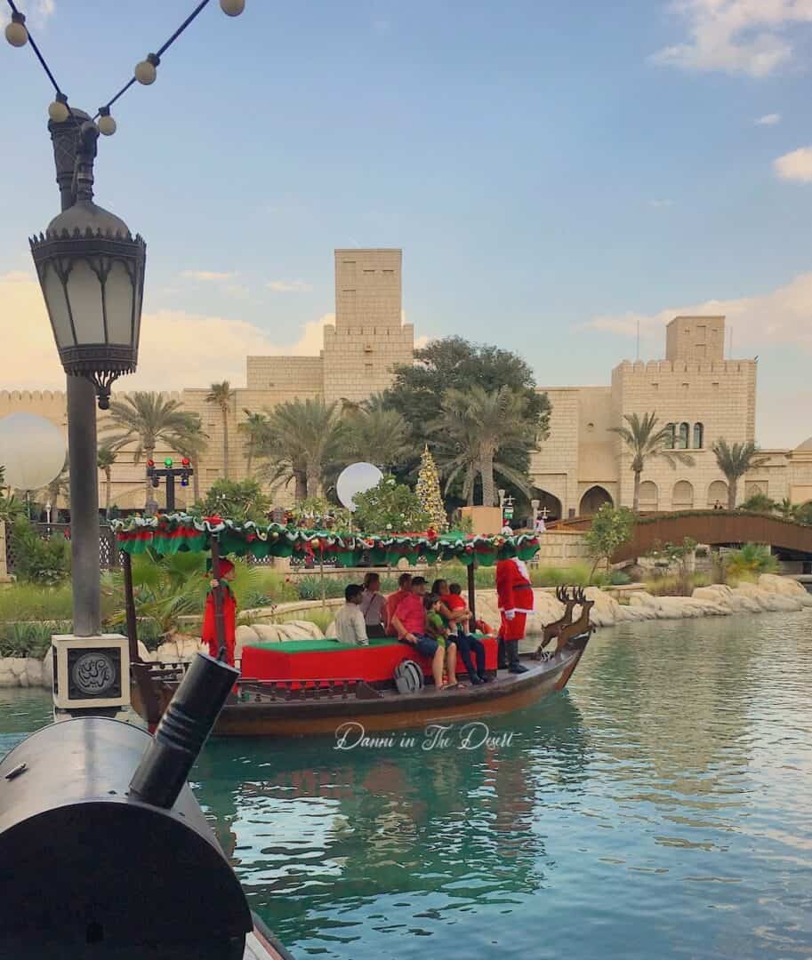 Madinat Jumeriah's abras decorated with Christmas decorations floating along the lakes with an elf and santa, with the Christmas markets in the background with their large Christmas tree