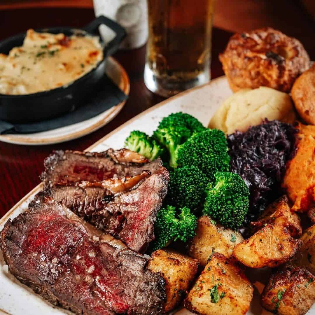 A traditional beef roast dinner with potatoes and vegetables along with cauliflower cheese as a side dish on a wooden tables at McGettingans