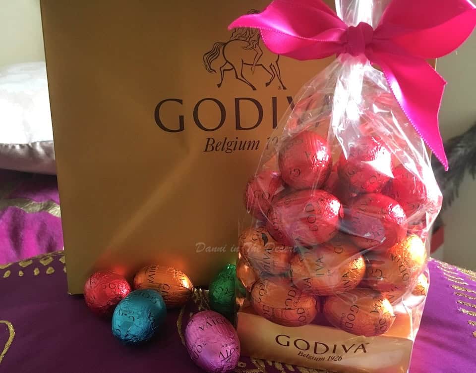 A bag of chocolate easter eggs with different colour foil wrappers, with some inside the bag and a few next to it and a Godiva chocolate gift bag behind