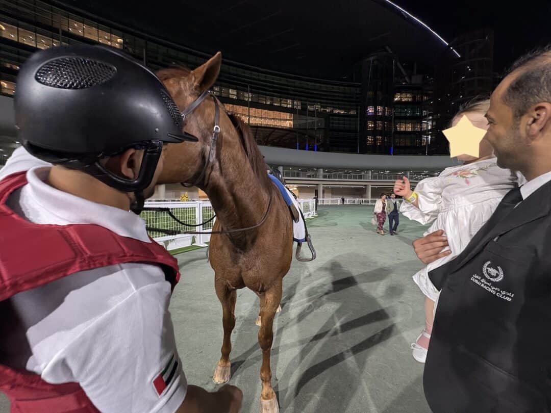 The manager of the restaurant at Dubai racing on the right in a black jacket, holding a toddler to see the thoroughbred racing horse with a ride on the right wearing a red and white racing uniform at Meydan Racing Course