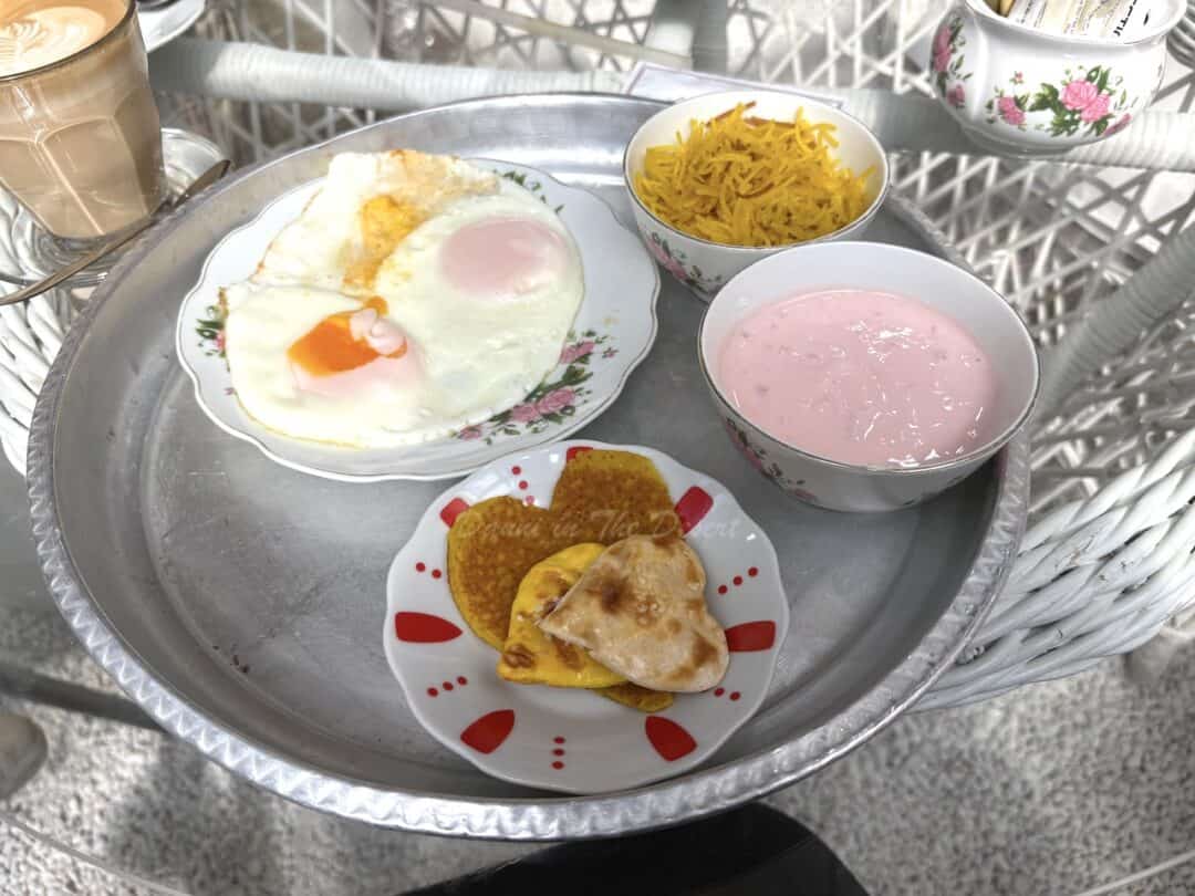 Two friend's eggs, sweet vermicelli noodles in a bowl, strawberry yoghurt and heart shaped breads with pancakes on a white and red plat. All served on a large silver tin tray.