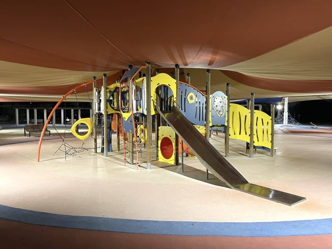 Blue and yellow outdoor play area with a metal slide, with a circus tent style cover over it at night time at Quran Park in Dubai