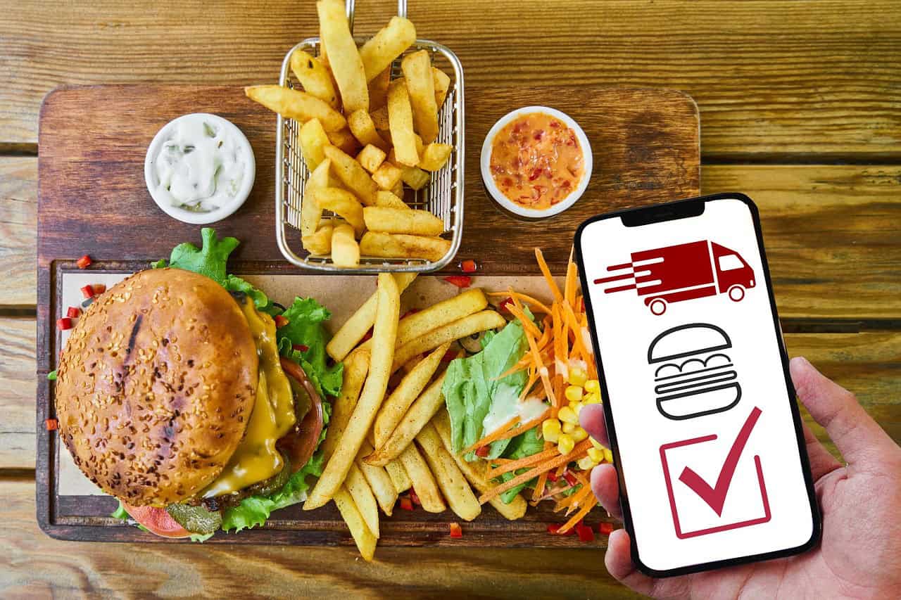 A food delivery app on an iPhone with a burger, fries and salad in the background representing food ordered online