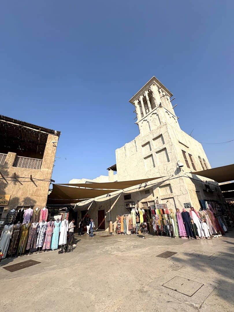 Fabrics and traditional Emirati outfits mixed with modern clothes on display in front of traditional Emirati historic houses with it's famous wind tower towering above