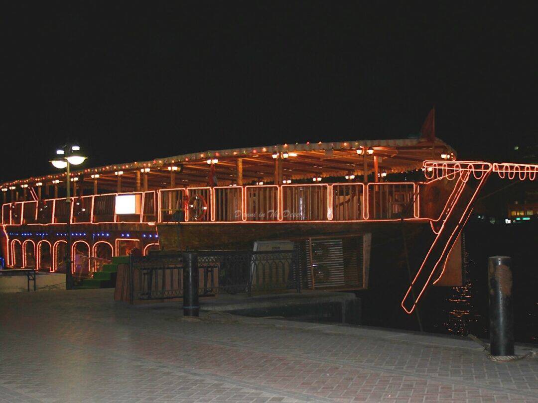 Large dhow boat docked in Dubai Creek with it's gold glowing neon lights around its wooden features during the night time before it sets sail