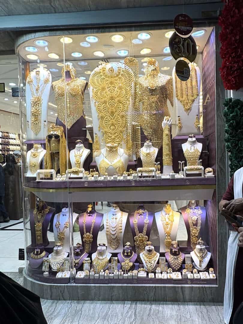 Various large, ornate 22 carat gold pieces of jewellery displayed on white neck mannequins with smaller pieces also displayed behind the glass in one of the shops in Dubai's gold souk