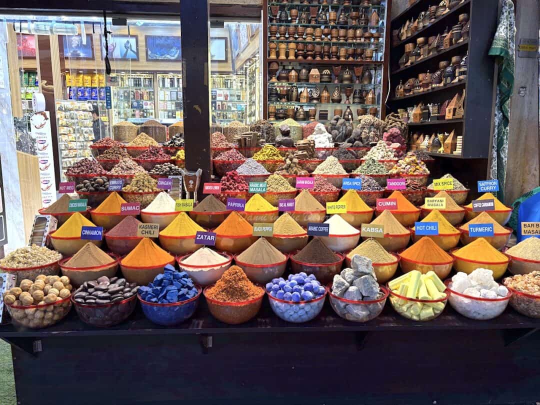 Various colourful spices pilled high in individual bowls and placed on layered shelves to show off the contents to prospective customers at the front of the shop in Dubai's spice souk with various items for sale in the background
