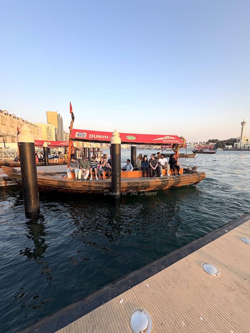 A traditional wooden abra with the classic Dubai red ETA canopy, docked at Deira Old Souq stop on Dubai creek as people sit on the boat waiting to leave