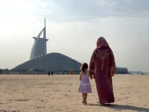 A mother and daughter, the daughter wearing a summer dress with a mother wearing a pink abaya walking towards Marsa Al Arab and Burj Al Arab on Umm Suqeim Beach