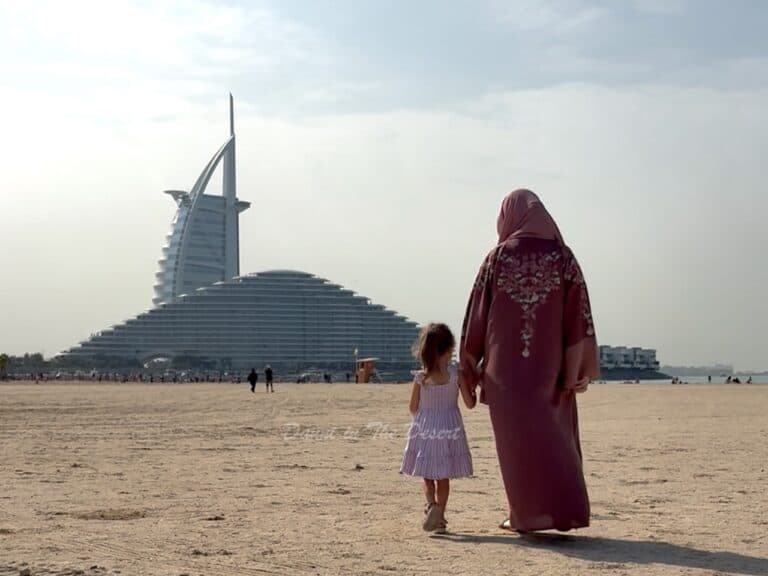 A mother and daughter, the daughter wearing a summer dress with a mother wearing a pink abaya walking towards Marsa Al Arab and Burj Al Arab on Umm Suqeim Beach